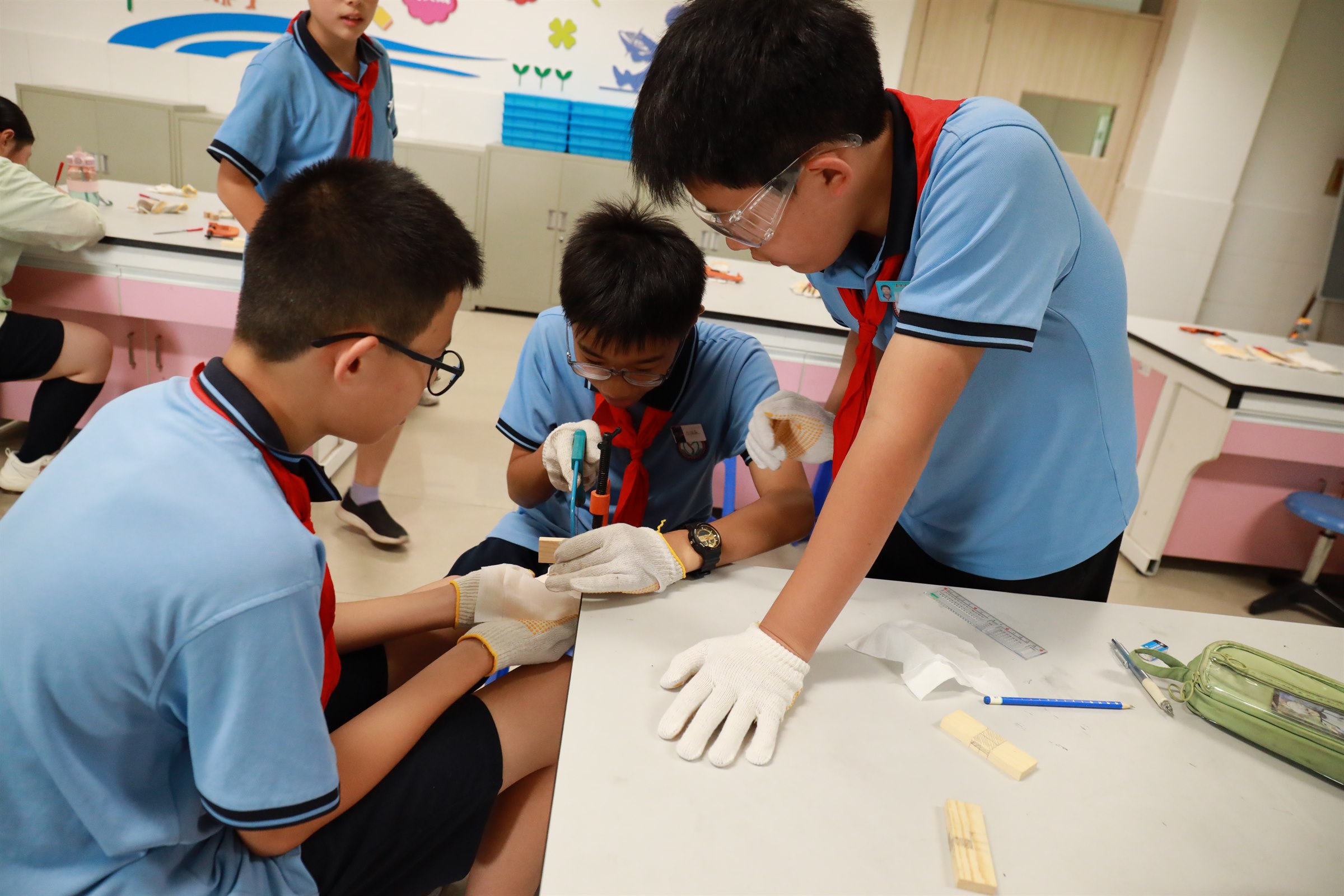 Three students use hand tools to work on a small wooden piece at a classroom table.